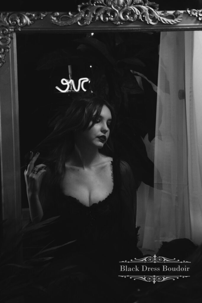 IMG_0125-bw A black and white photo of a woman in a dark lace dress looking into a mirror, hand in hair, with a neon sign above reflecting in the glass.