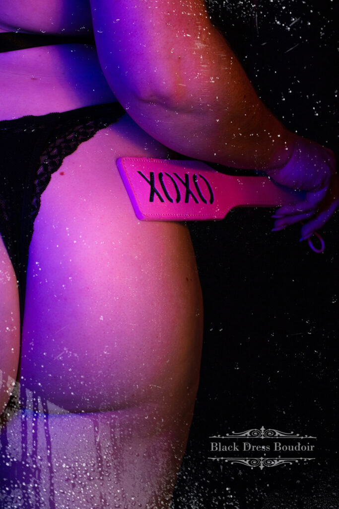 A close-up of a woman holding a pink paddle with "xoxo" against her buttock with water droplets.