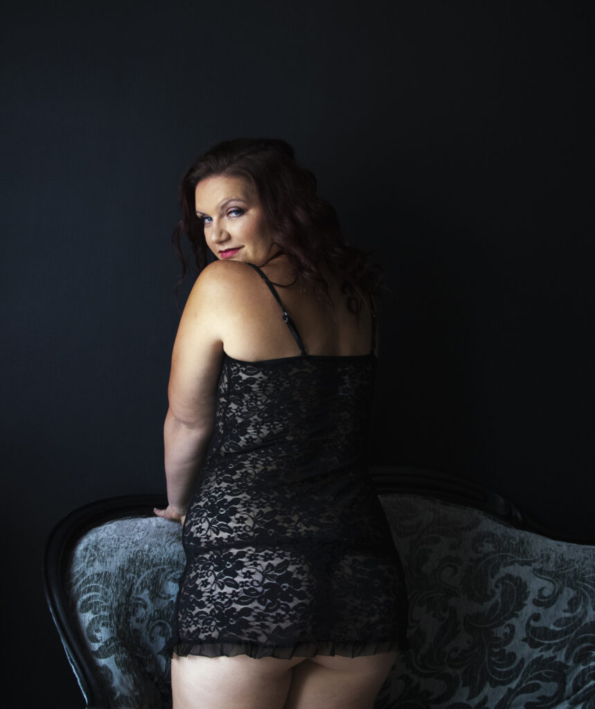 a Woman in black lace chemise kneeling on a couch looking back over her shoulder towards the viewer