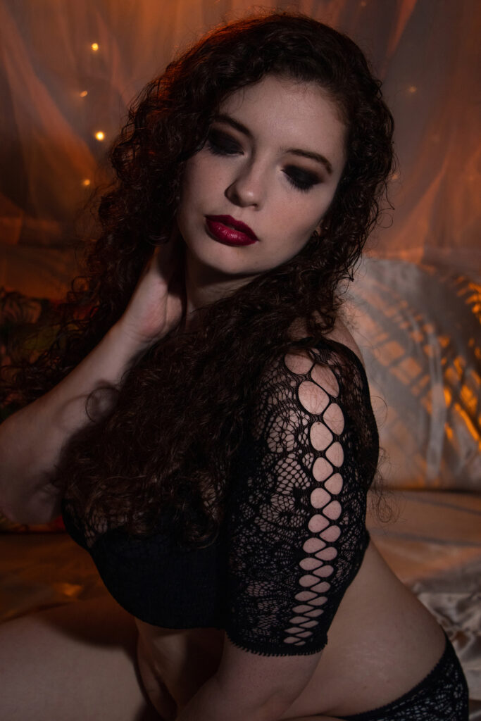 a A woman in full coverage black cut-out lingerie posing on a satin sheet bed wearing stunning red lipstick