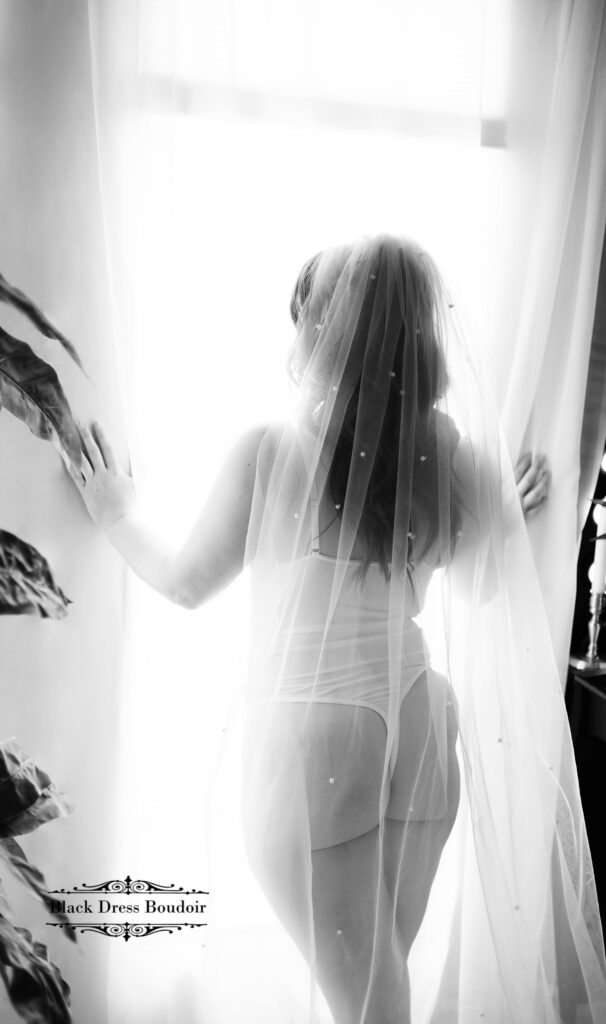 A black and white Flint MI bridal boudoir photo of a woman standing in front of a large window a pearl view draped down her back with large plants flanking her