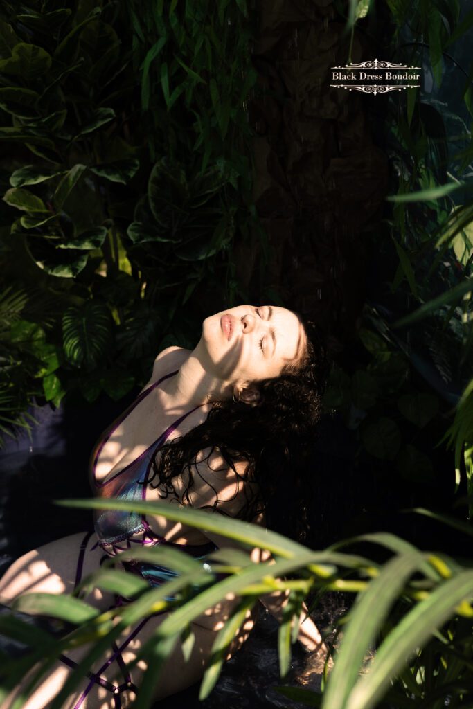 A woman in shimmering boudoir lingerie gracefully leans back, basking in dappled sunlight. Lush greenery surrounds her, creating an ethereal, serene atmosphere.
