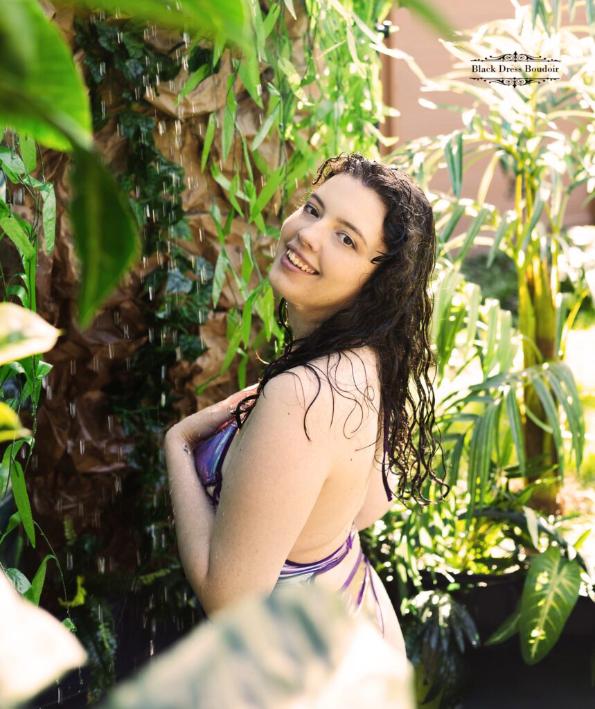Smiling woman with wet hair, in a swimsuit, standing under a cascade of water surrounded by lush green plants, exuding joy and relaxation at a clio, michigan boudoir studio
