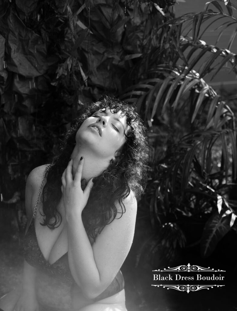 A mini boudoir portrait of a woman with curly hair leans back with closed eyes, her hand gently touching her neck. Surrounded by lush plants in soft lighting in Clio, Michigan