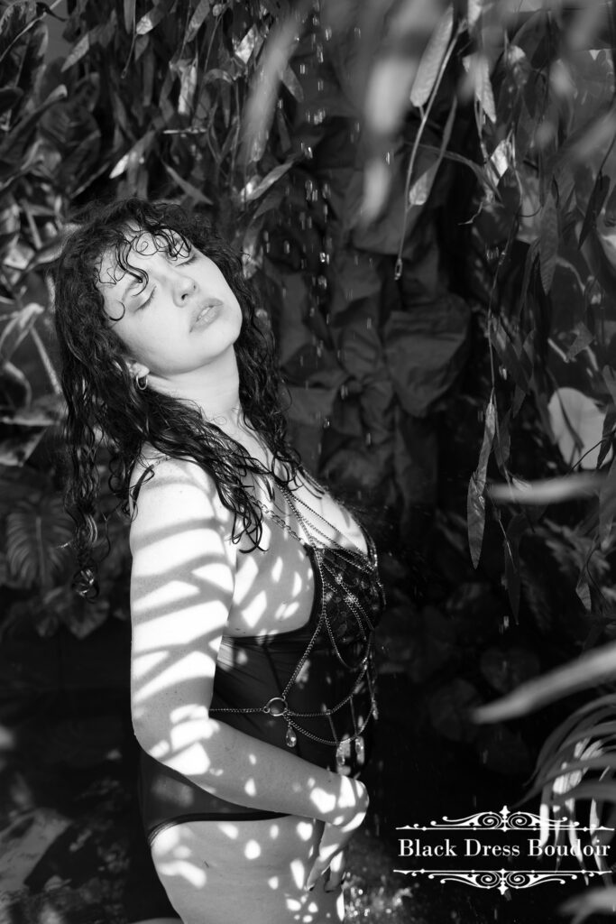 Woman in a floral bodysuit stands under a waterfall, eyes closed, surrounded by lush foliage at her michigan boudoir mini session