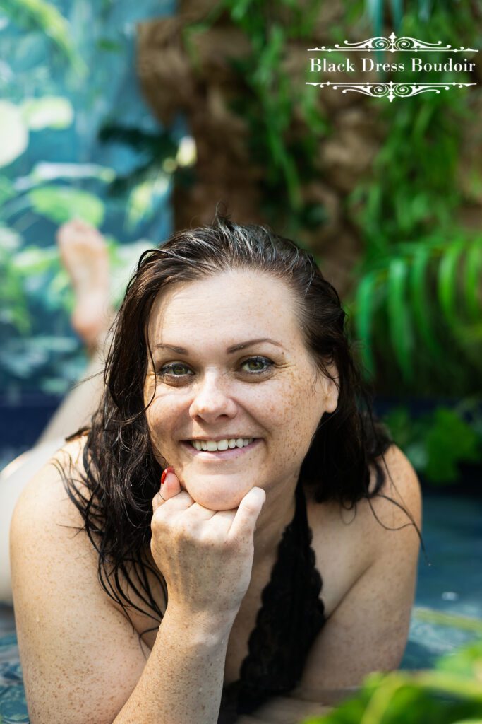 Photograph of a Michigan at a Mid-Michigan boudoir studio of a woman with wet hair smiling in a pool, surrounded by lush greenery. Her hand rests on her chin, conveying a relaxed, joyful mood.