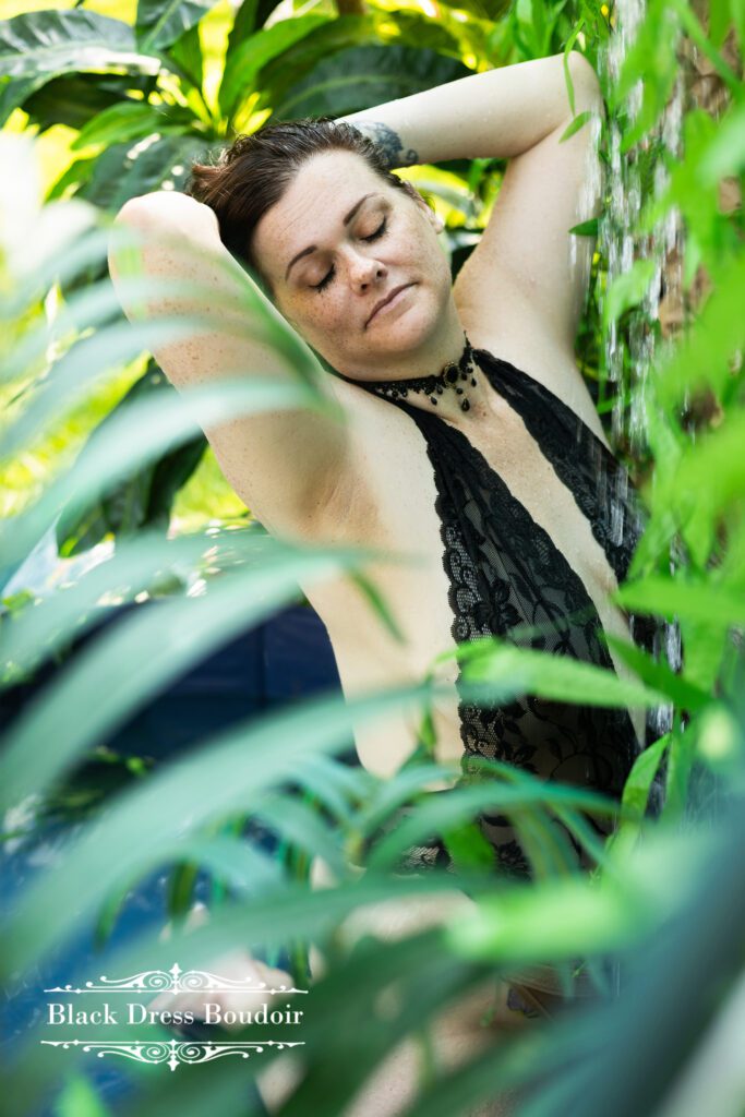Woman in black lace boudoir lingerie stands among lush green plants, eyes closed, arms raised, appearing relaxed and serene. Sunlight filters through leaves in Mid Michigan